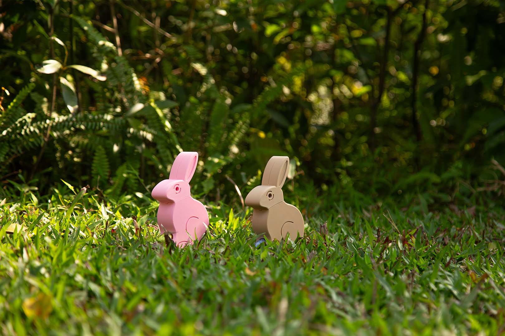 Two 3D-printed Easter Egg Dispenser Bunnies, one pink and one beige, sit on a grassy field with trees in the background, ready to dispense colorful chocolate eggs.