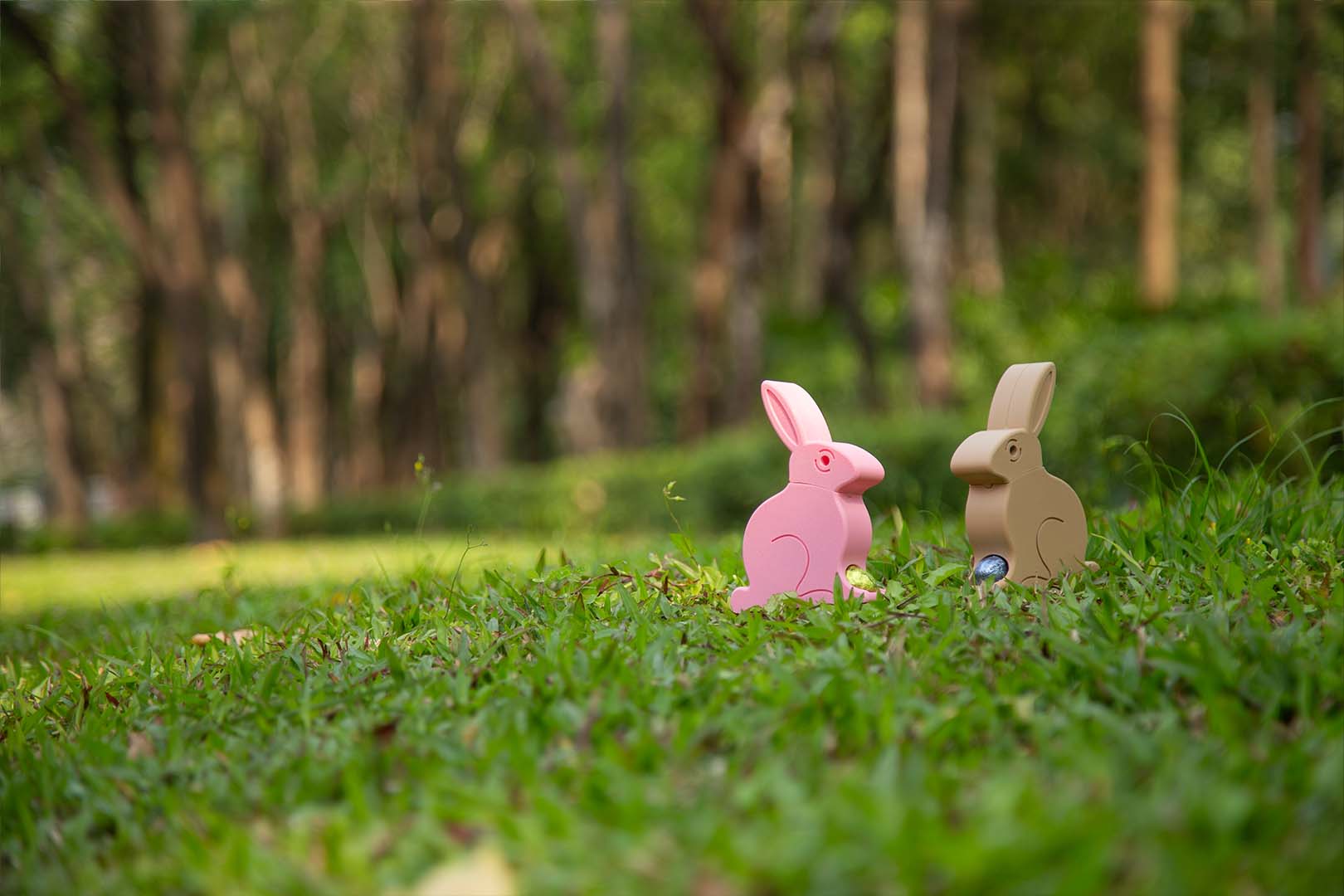 A pink 3D-printed Easter Egg Dispenser Bunny next to a beige one on a grassy field, with a blue and a yellow chocolate egg placed in front of them.