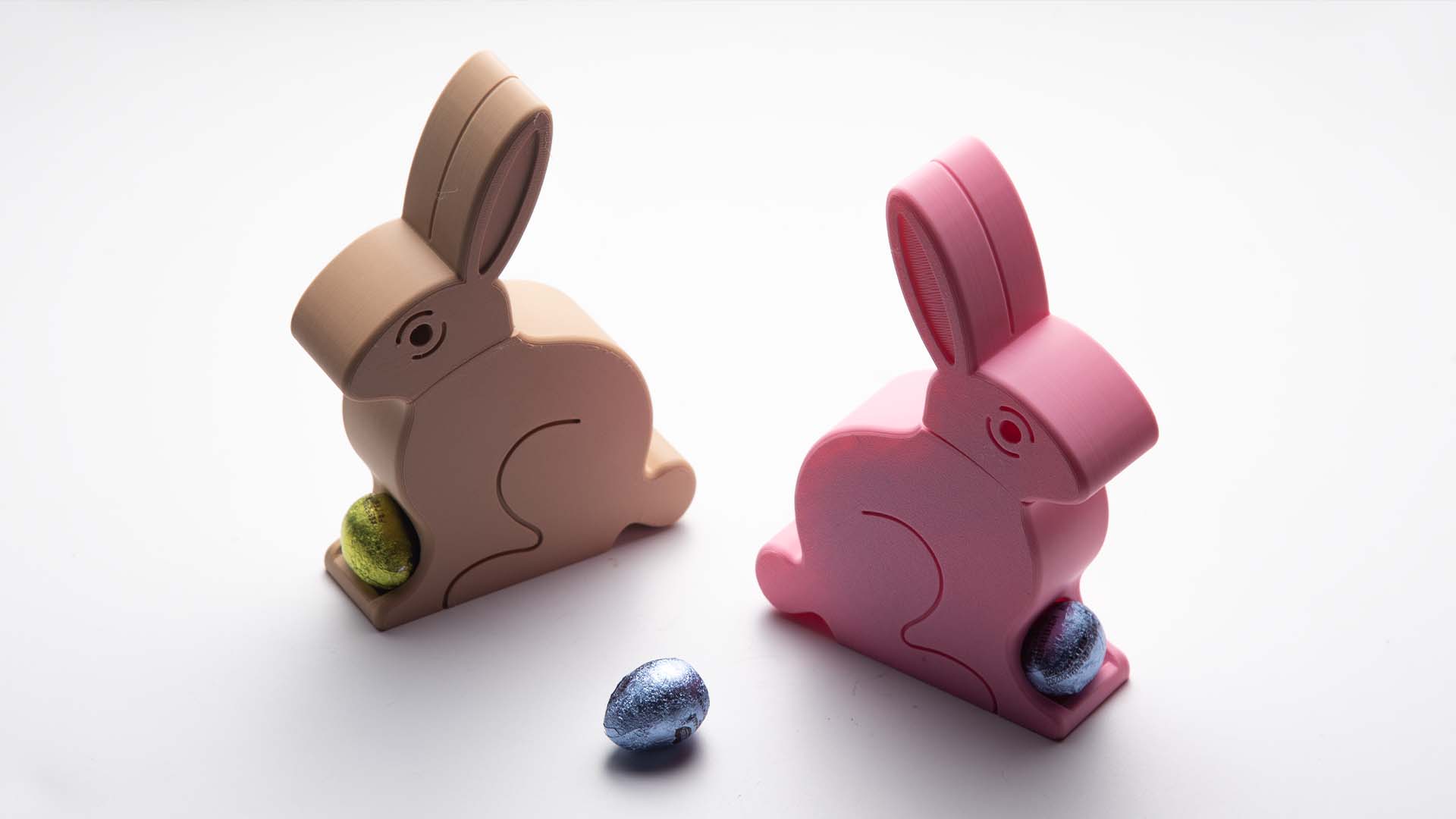 A beige and a pink 3D-printed Easter Egg Dispenser Bunny on grass, with the pink bunny dispensing a yellow egg and a blue egg nearby.