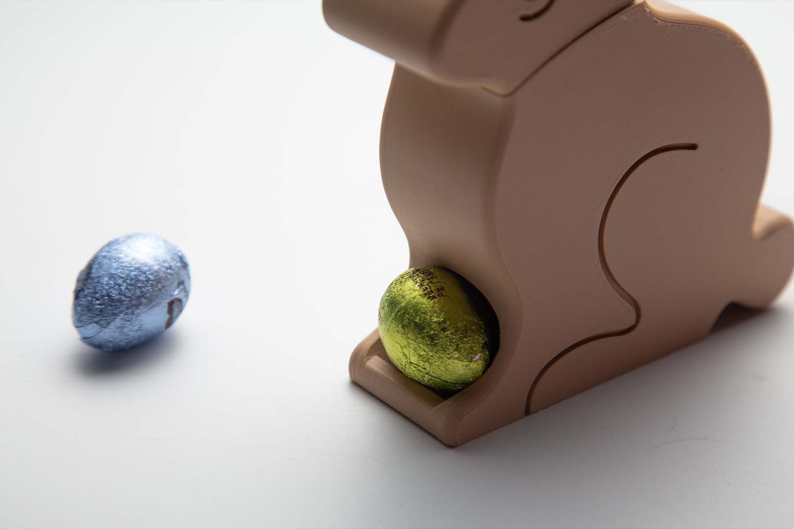 A pink 3D-printed Easter Egg Dispenser Bunny next to a beige one on a white background, with a blue egg in front of the pink bunny.