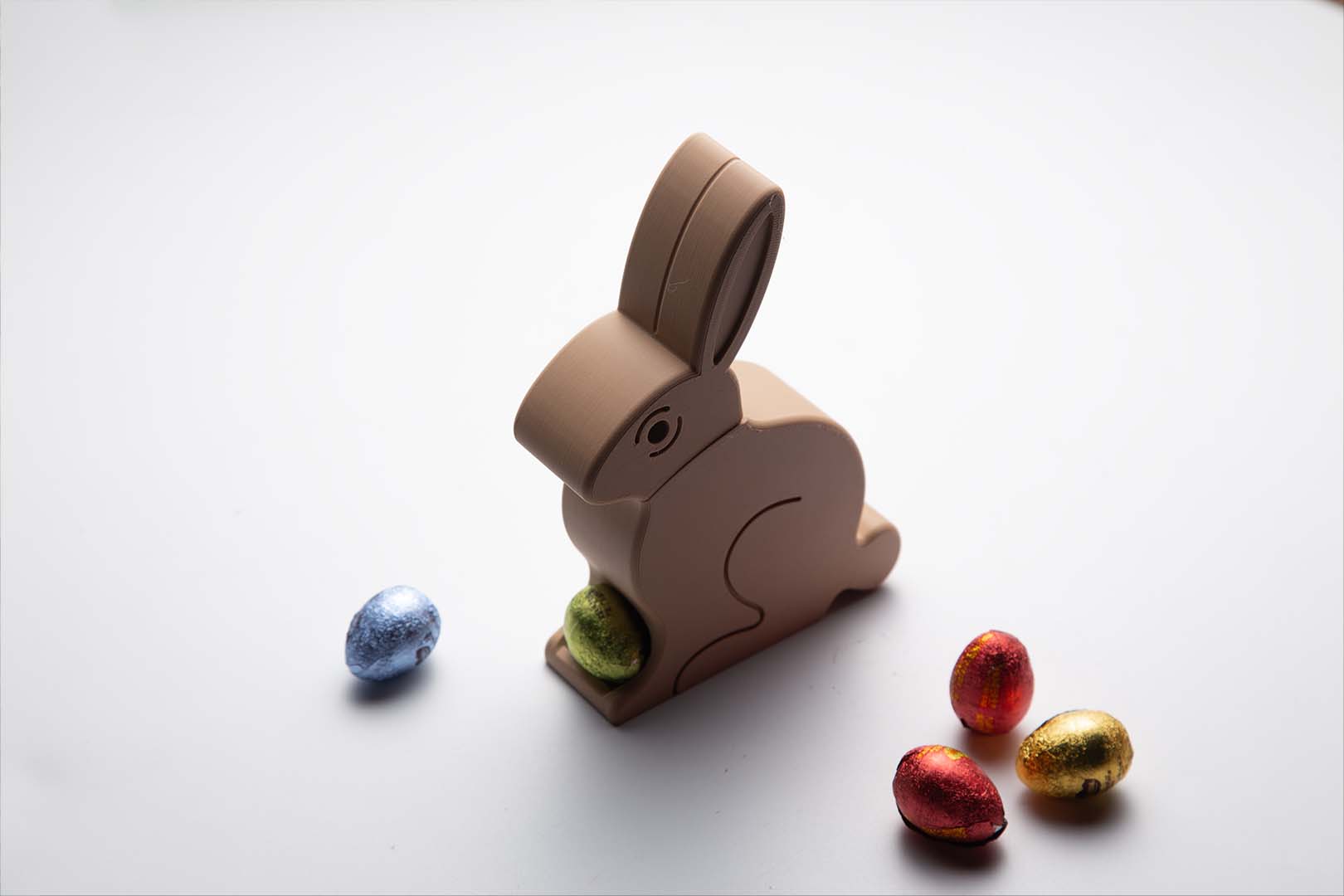 A beige 3D-printed Easter Egg Dispenser Bunny on a white background, dispensing a yellow egg with a blue egg nearby.