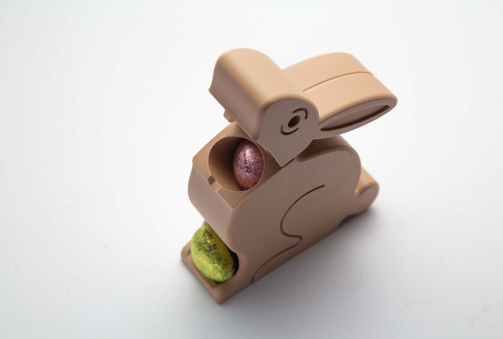 A beige 3D-printed Easter Egg Dispenser Bunny on a white background, with a yellow egg being dispensed and blue, red, and gold eggs scattered around it.